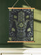 Botanical Hamsa wall tapestry featuring intricate sage green and lavender design on black background, showing a protective hand symbol surrounded by leaf motifs and floral elements, hanging from wooden dowels with natural twine.